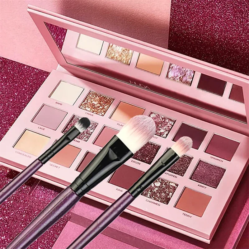 Close-up view of a pink eyeshadow palette with three makeup brushes resting on top. The palette is open, showcasing various shades of pink, brown, and glitter eyeshadows. The brushes are purple and black, with soft bristles.