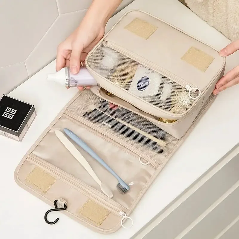 A beige travel cosmetic bag is shown open, revealing its various compartments filled with toiletries and makeup items. The bag is laid on a white countertop, with a person's hands visible in the upper part of the image, suggesting they are organizing the contents. A small, black box is visible in the bottom left corner.