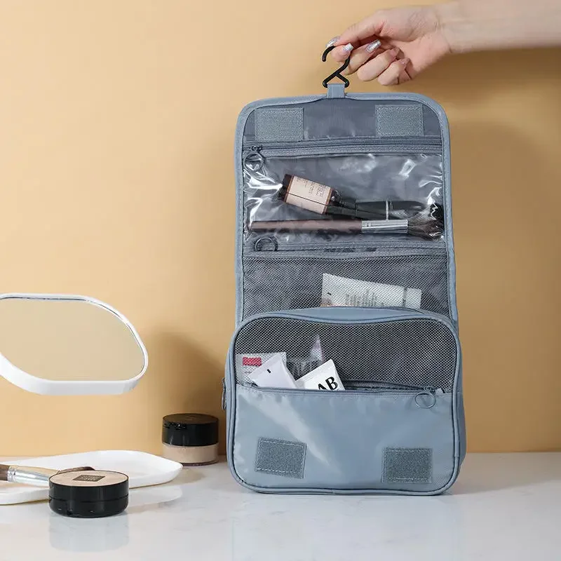 A light gray hanging travel makeup bag is shown open, revealing its various compartments. The bag is hung from a hook held by a person's hand. The bag contains various cosmetics and toiletries. A mirror and other cosmetic containers are on a table in front of it.