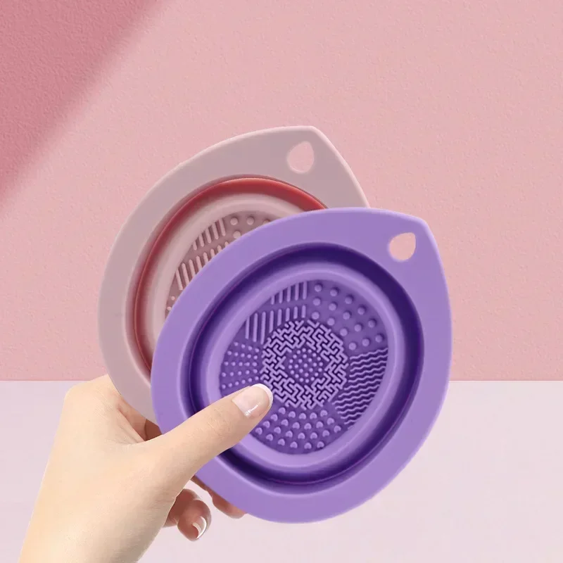 Two silicone cleansing pads in purple and beige are held in a hand against a pink background.