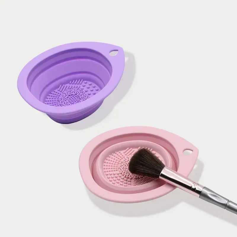 Two silicone makeup brush cleaning bowls, one purple and one pink, with a makeup brush resting in the pink bowl.