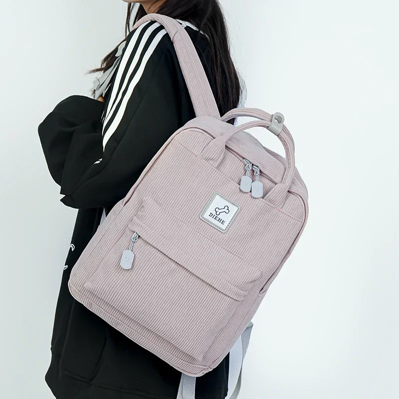 A light pink corduroy backpack with a small bone-shaped logo. The backpack is being worn by a person wearing a black and white striped sweater.
