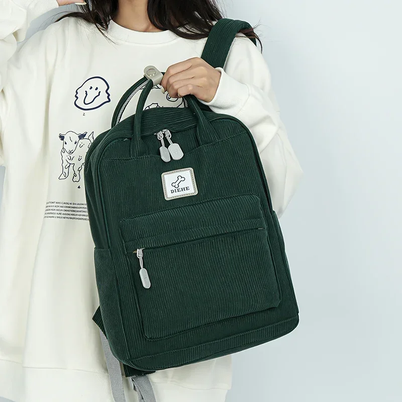 A dark green corduroy backpack with light gray accents. The backpack has a top handle and adjustable shoulder straps. A woman is holding the backpack in her hands. The background is a light gray.