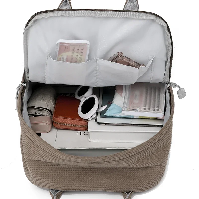 A taupe-colored corduroy backpack is open to reveal its contents: a gray interior with multiple pockets, a compact umbrella, sunglasses, a wallet, a tablet, and other personal items.