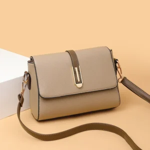A beige crossbody bag with a gold clasp sits on a beige surface.