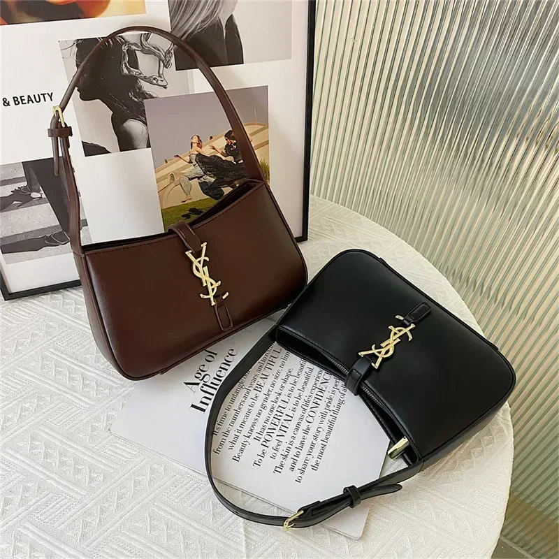 Two stylish shoulder bags, one brown and one black, rest on a white surface with a magazine. Both bags feature a gold YSL-style logo.