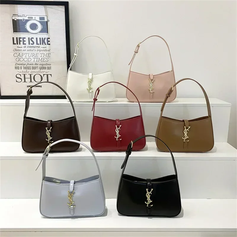 Six designer handbags in a row on a white shelf. Each bag is a different color and is a half-moon shape. The bags are arranged from left to right: white, pink, brown, red, light gray, and black. Each bag features a prominent gold Yves Saint Laurent logo.