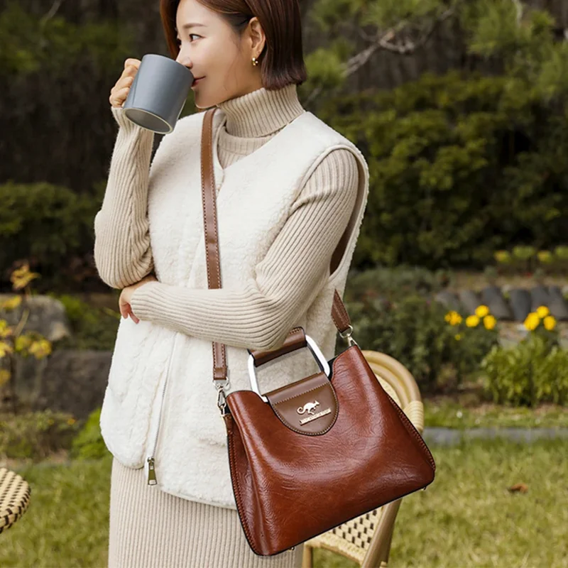 A woman in a cream-colored sweater vest holds a dark brown leather handbag.