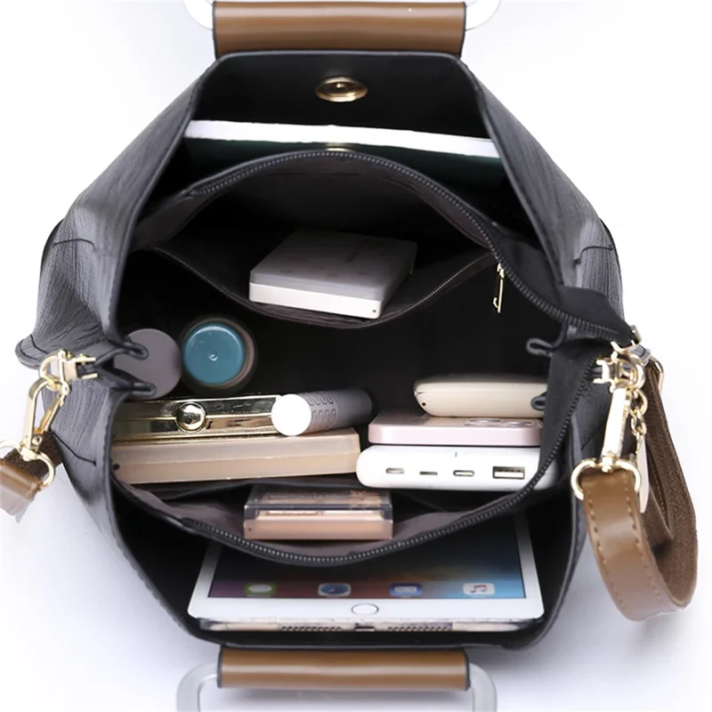 A black handbag with brown handles is open, revealing its well-organized interior filled with various items such as a tablet, makeup, and other personal belongings. The bag's spacious design and multiple compartments are clearly visible.