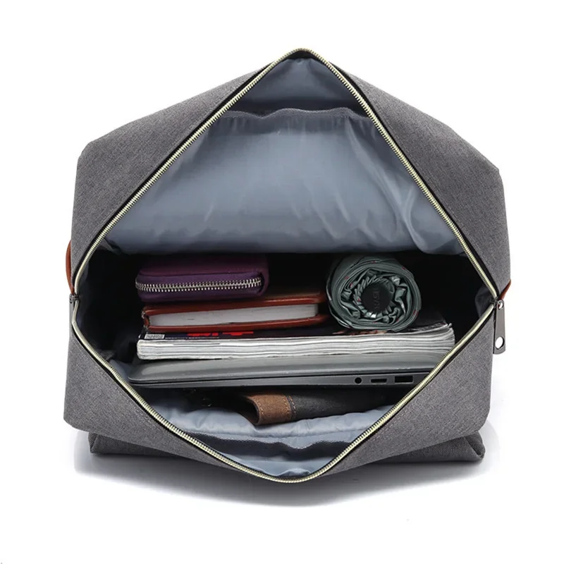 A gray canvas backpack is open, revealing its interior and the various items it contains. The backpack is lined with a light gray fabric. The items inside include a purple wallet, notebooks, magazines, a laptop, and a folded umbrella. The backpack's zipper is gold-colored, and the pull tabs are made of brown leather. The background is white.