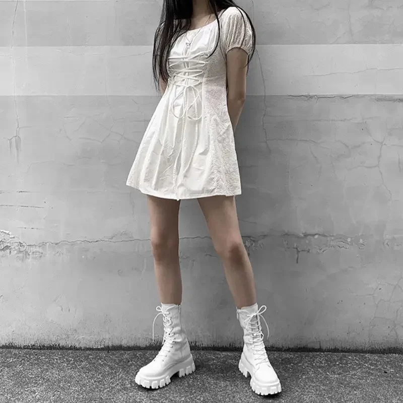 A person wearing a white dress and white boots stands in front of a gray wall.
