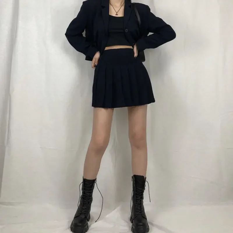 A person wearing a black blazer, black pleated skirt, and black combat boots. The person is standing in front of a white wall.