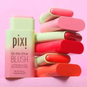 A close-up shot of a stack of Pixi On-the-Glow Blush sticks in various shades of pink, orange, and red. The packaging is pastel green and the brand name is prominently displayed on the front of one stick.