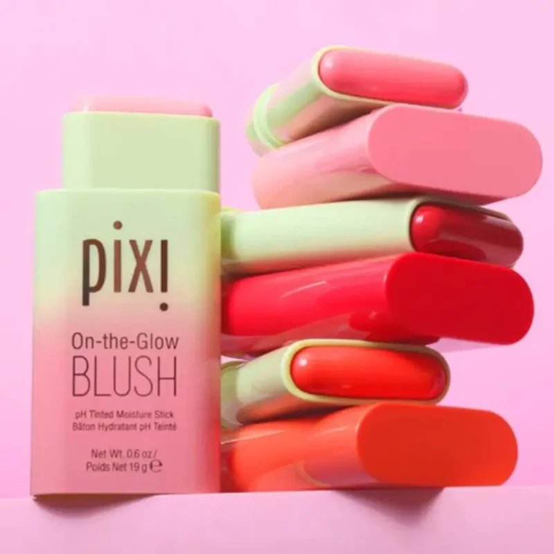 A close-up shot of a stack of Pixi On-the-Glow Blush sticks in various shades of pink, orange, and red. The packaging is light green and the text on the product reads 'pixi', 'On-the-Glow', 'BLUSH', 'pH Tinted Moisture Stick', and other details.
