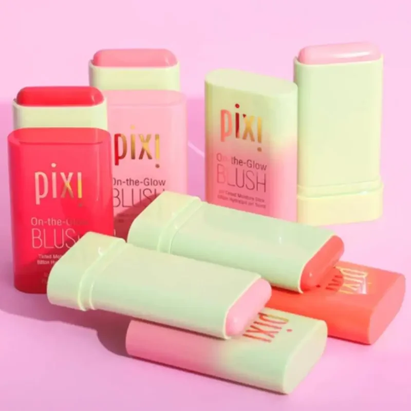 Close-up view of several Pixi On-the-Glow Blush sticks in various shades, arranged on a pink surface. The packaging is pastel green and pink, with the Pixi logo prominently displayed.