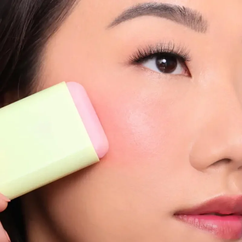 Close-up view of a woman applying a pastel pink and green cream blush stick to her cheek. Her skin is smooth and makeup is minimal.