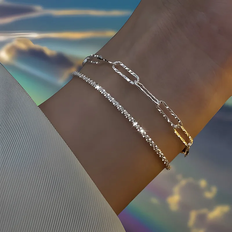 Close-up of a woman's wrist wearing two delicate bracelets. One bracelet has a sparkly, thin chain, while the other features a chain of interconnected rectangular links. The background is blurred and features a vibrant sky with clouds and a hint of a rainbow.