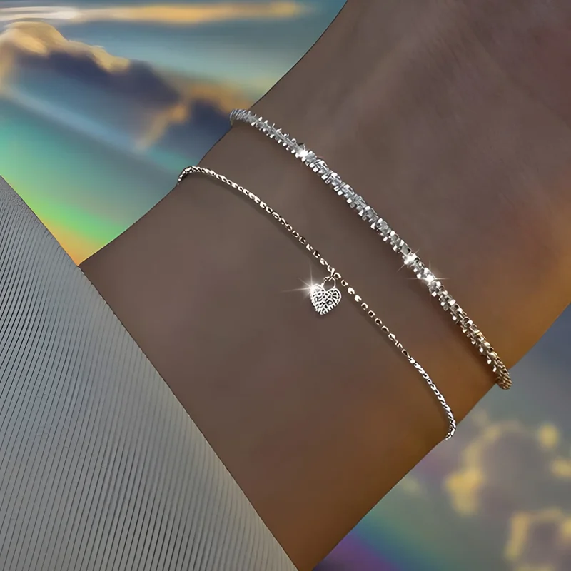 Close-up of a woman's wrist adorned with two delicate silver bracelets. One bracelet features a charming heart-shaped pendant.