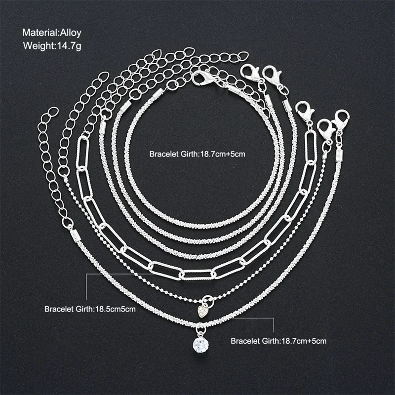 Seven layered silver anklets with varying chain styles and a small heart charm with a clear stone pendant.