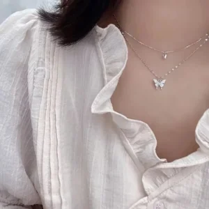 Close-up of a woman wearing a delicate layered necklace with butterfly pendants. The necklace is made of thin gold chains and features small, sparkly butterfly charms. The woman is wearing a light beige, ruffled blouse.