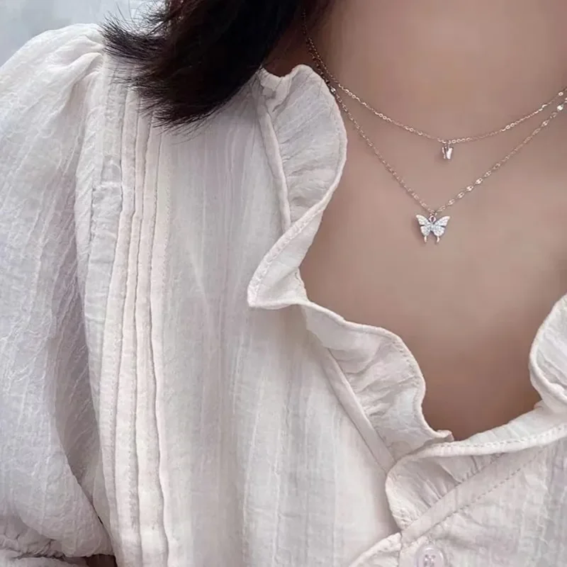 Close-up of a woman wearing a delicate layered necklace with butterfly pendants. The necklace is made of thin gold chains and features small, sparkly butterfly charms. The woman is wearing a light beige, ruffled blouse.