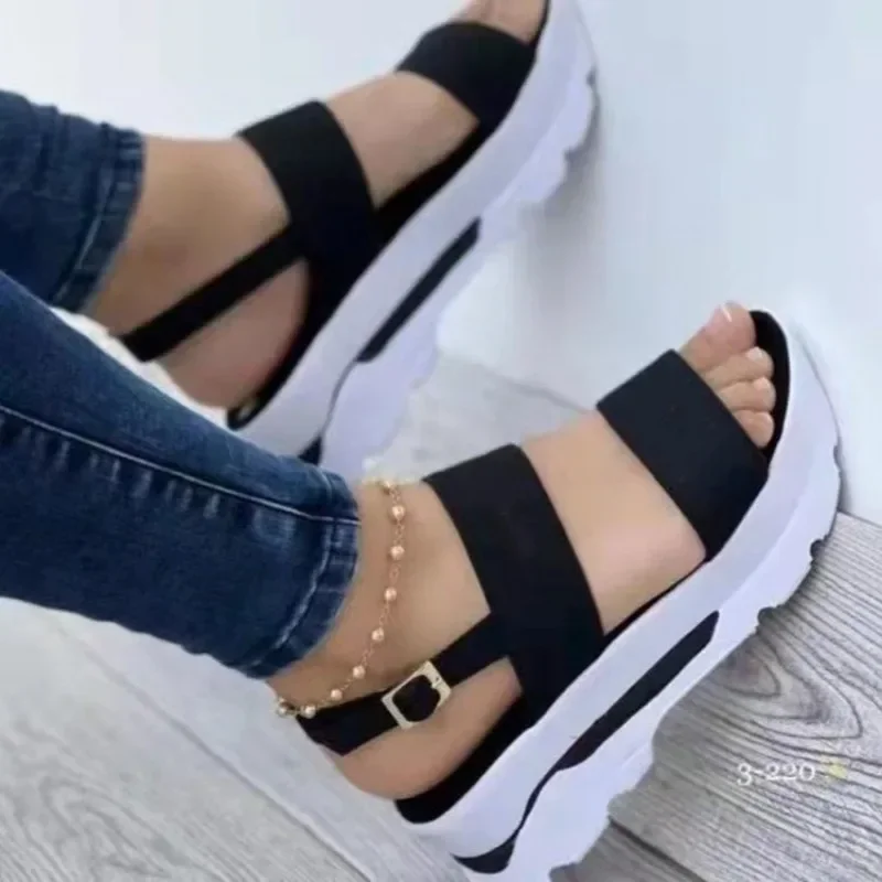 A close-up shot of a woman's feet wearing a pair of trendy black and white platform sandals. The sandals have two straps across the top of the foot and a chunky white platform sole. The woman is wearing dark blue jeans. The background is a light-colored wall.
