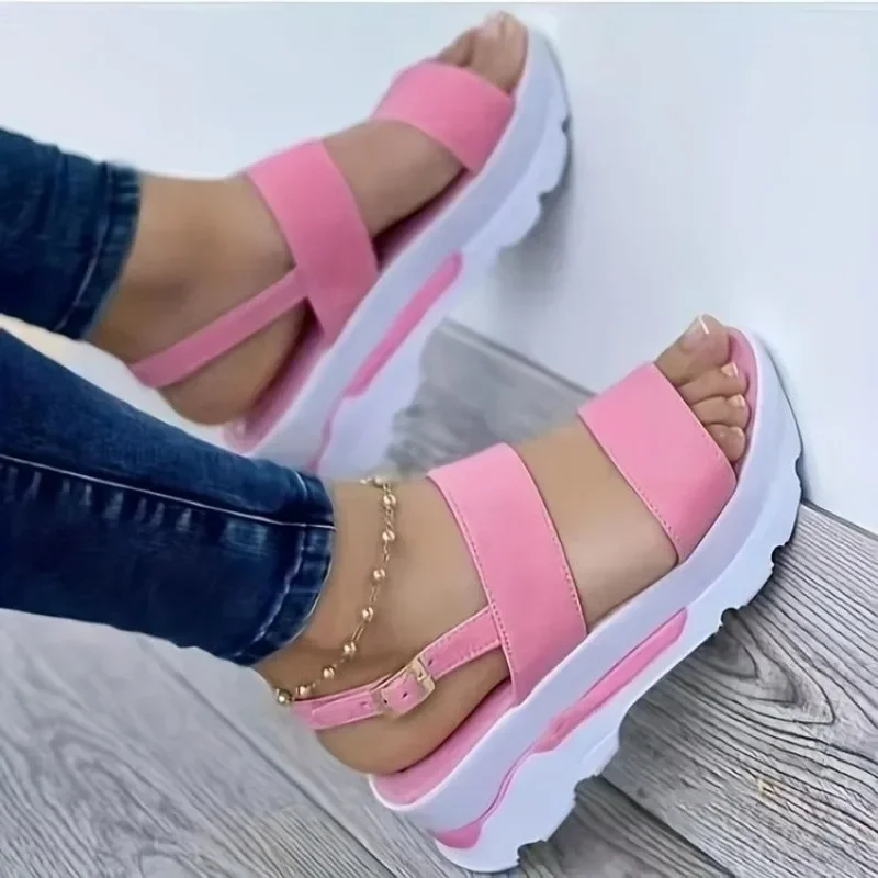 A pair of pink platform sandals with white soles. The sandals are worn by a person with their legs crossed. The background is a light-colored wooden floor.