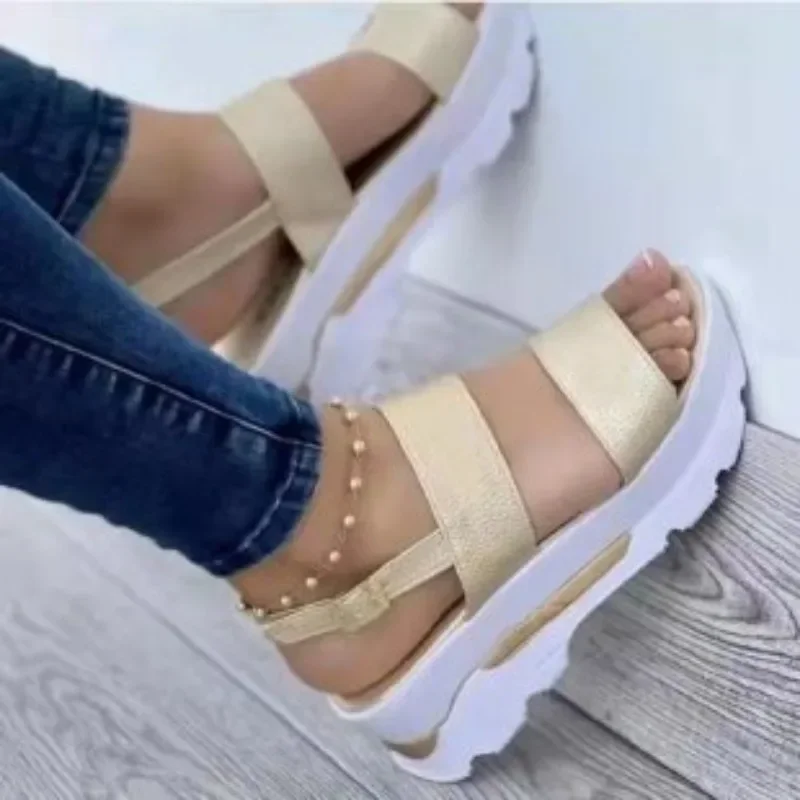 Close-up image of a person's legs wearing a pair of gold platform sandals. The sandals have white soles and are worn with blue jeans.