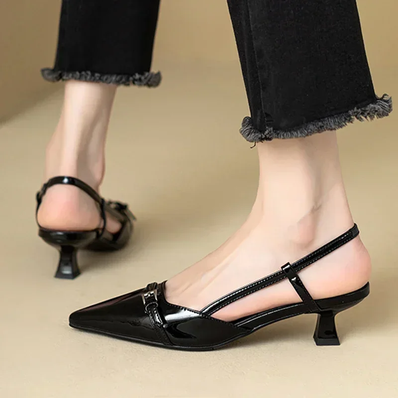 A woman's feet wearing black patent leather kitten heels with ankle straps. The shoes are pointed-toe and have a small buckle detail. The woman is wearing black jeans.