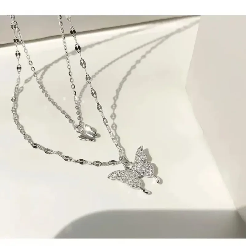 A close-up shot of a delicate silver necklace with a sparkling butterfly pendant. The necklace is layered, with a smaller butterfly charm near the top. The background is a clean, white surface.