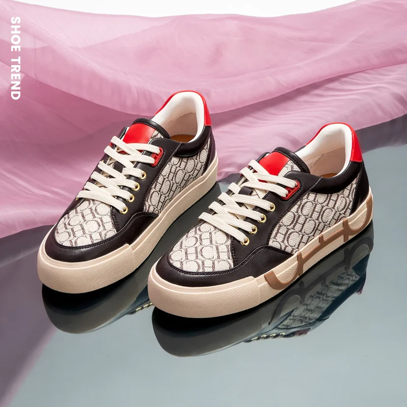 A pair of stylish brown and beige sneakers with red accents sits on a pink fabric background. The sneakers have a cream-colored sole and a monogram pattern on the upper.