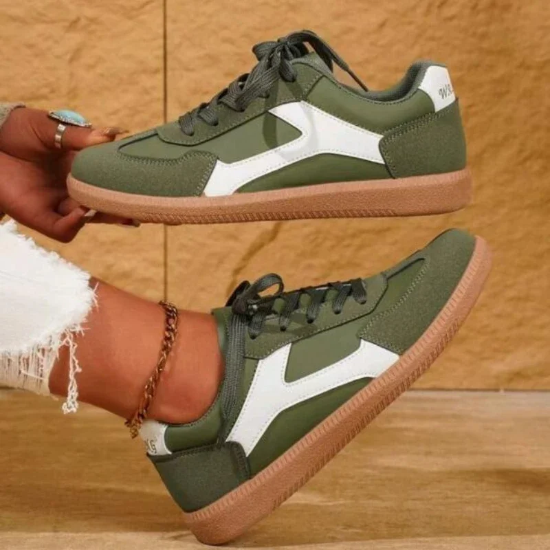A pair of olive green and white sneakers with a tan sole. The sneakers are shown on a person's feet, and the background is a light brown wooden surface.