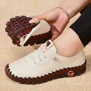 A close-up shot of a woman's hand holding up a pair of off-white leather shoes with a burgundy sole. The shoes have a stitched pattern around the edge and a small logo on the side. The woman is wearing a simple bracelet.