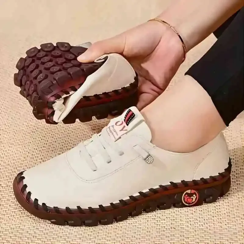 Close-up of a person's feet wearing a pair of off-white casual shoes with a unique textured sole. The shoes are being held in the person's hand, which is adorned with a bracelet. The shoes appear to be made of leather and have a comfortable design.