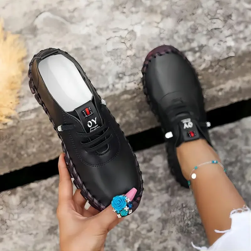 Close-up view of a person holding a black leather shoe with decorative stitching. Another black shoe is visible on the person's other foot. The shoes appear to be stylish and casual.