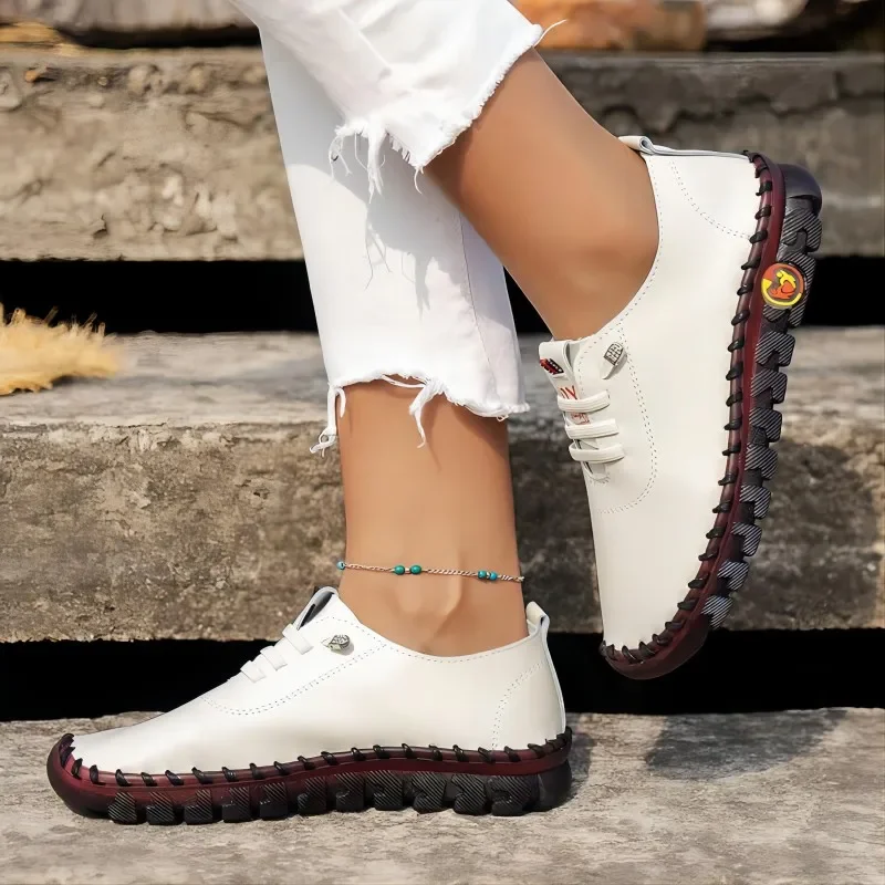 Close-up view of a woman's feet wearing stylish white leather casual shoes with a unique textured sole. The shoes are laced up and have a slightly rugged appearance. The woman is wearing white jeans that are slightly cuffed at the ankles. She is standing on some stone steps.