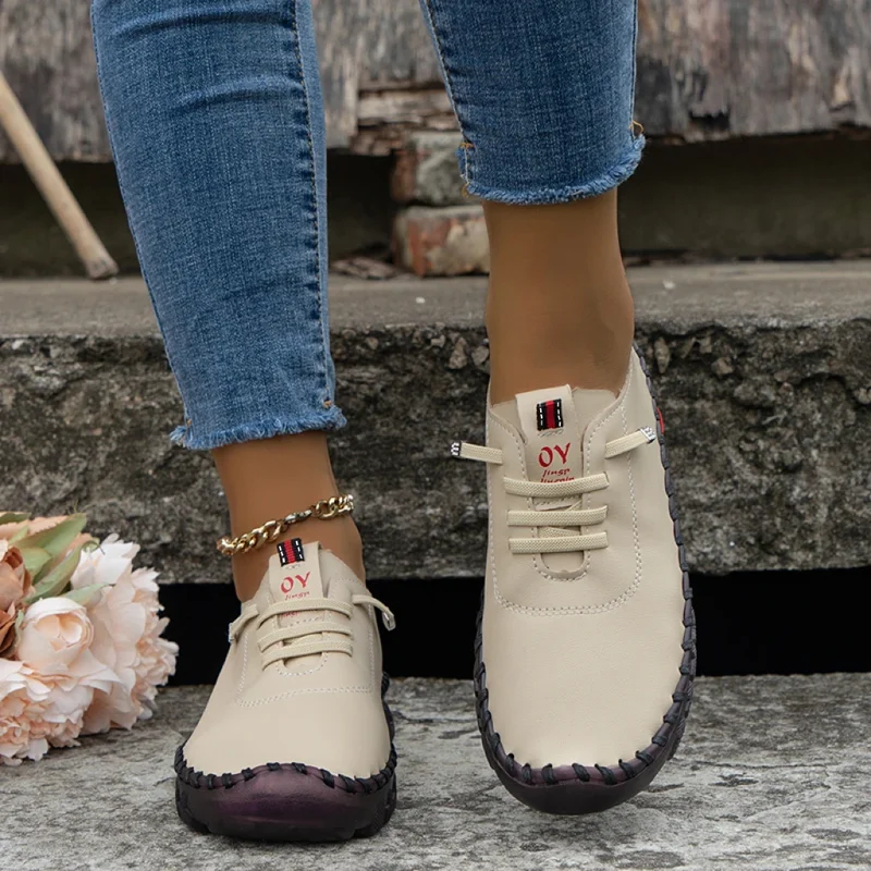 Close-up of a woman's feet wearing beige lace-up shoes with a dark brown stitching detail. The shoes are hand-stitched and have a casual style. The woman is wearing jeans that are rolled up at the ankles, and she is standing on a stone step.