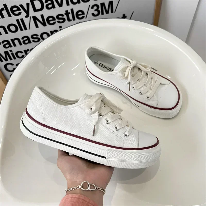 A pair of white canvas sneakers with maroon stripes sits in a white bowl. One shoe is slightly angled toward the viewer, and the other is positioned at a slight angle to the side. The shoes are simple in design with laces. The background is blurred but features some text and a muted pastel palette.
