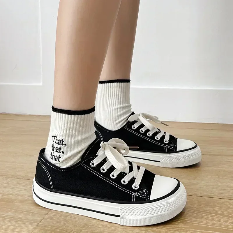 A pair of black canvas sneakers with white soles and laces. The sneakers are being worn by a person wearing white socks with black trim and the words "That, that, that" embroidered on them. The sneakers are sitting on a light brown wooden floor.