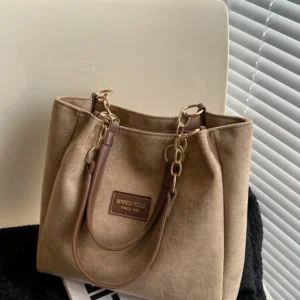 A light brown tote bag with gold chain straps sits on a white surface next to a black sweater. The bag is made of suede and has a Marco Polo logo.
