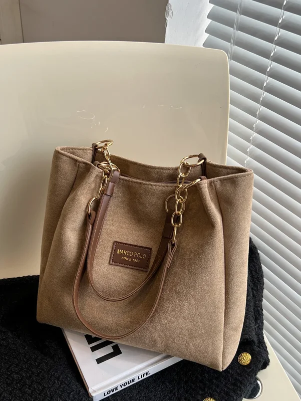A light brown tote bag with gold chain straps sits on a white surface next to a black sweater. The bag is made of suede and has a Marco Polo logo.