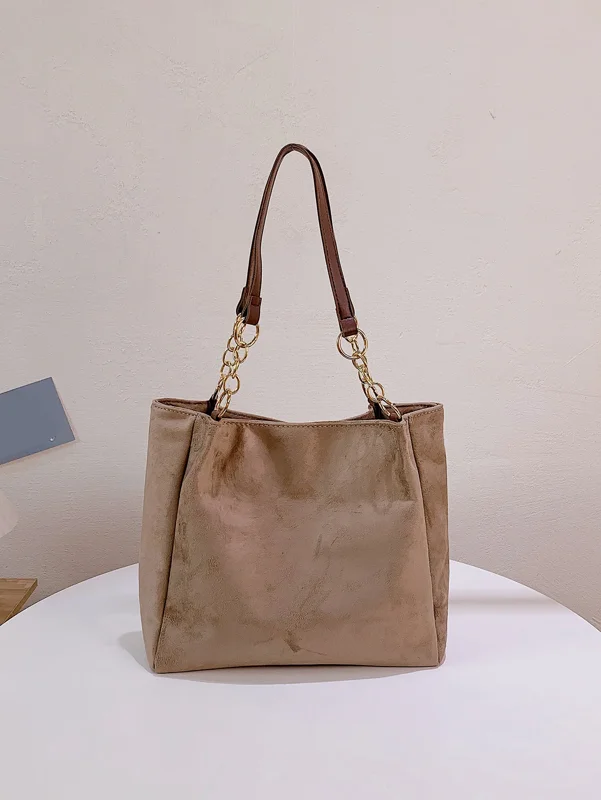 A beige tote bag with brown leather straps and gold chain details sits on a white table against a white wall.