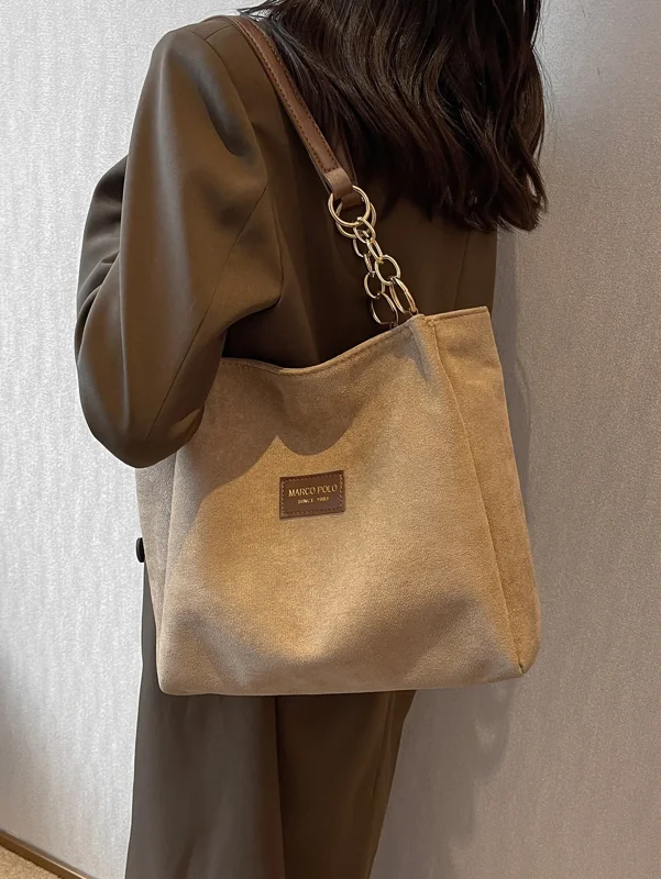 A woman in a brown blazer carries a beige tote bag with a brown leather strap and gold chain detail. The bag has a small brown leather tag with the words "MARCO POLO SINCE 1983" embossed on it.