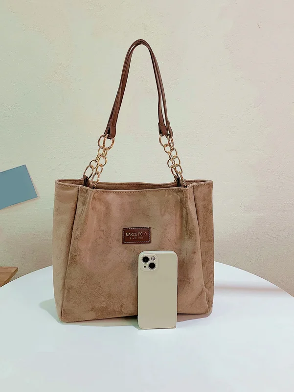 A taupe-colored suede tote bag with brown leather handles and gold chain accents sits on a white table. A cream-colored iPhone rests in front of the bag.