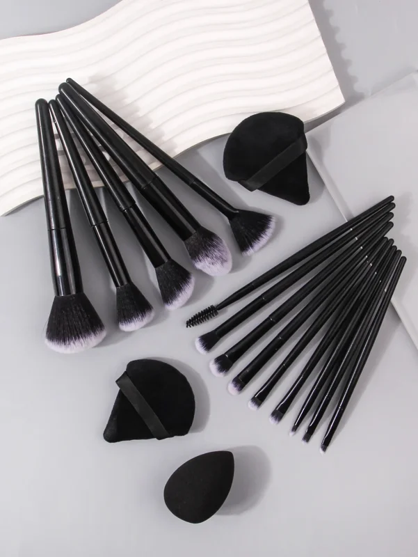 A set of black makeup brushes and sponges are arranged on a gray surface. The brushes are of various sizes and shapes, and the sponges are also different sizes. The background is a white wavy surface.