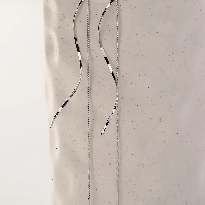 Close-up view of two long, thin silver chains hanging against a textured off-white background. The chains are slightly wavy and appear delicate. The background has a speckled appearance.