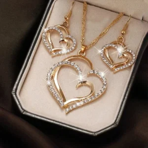 Close-up view of a gold-toned jewelry set featuring double heart pendants and earrings, adorned with sparkling crystals. The set is displayed in a beige box.