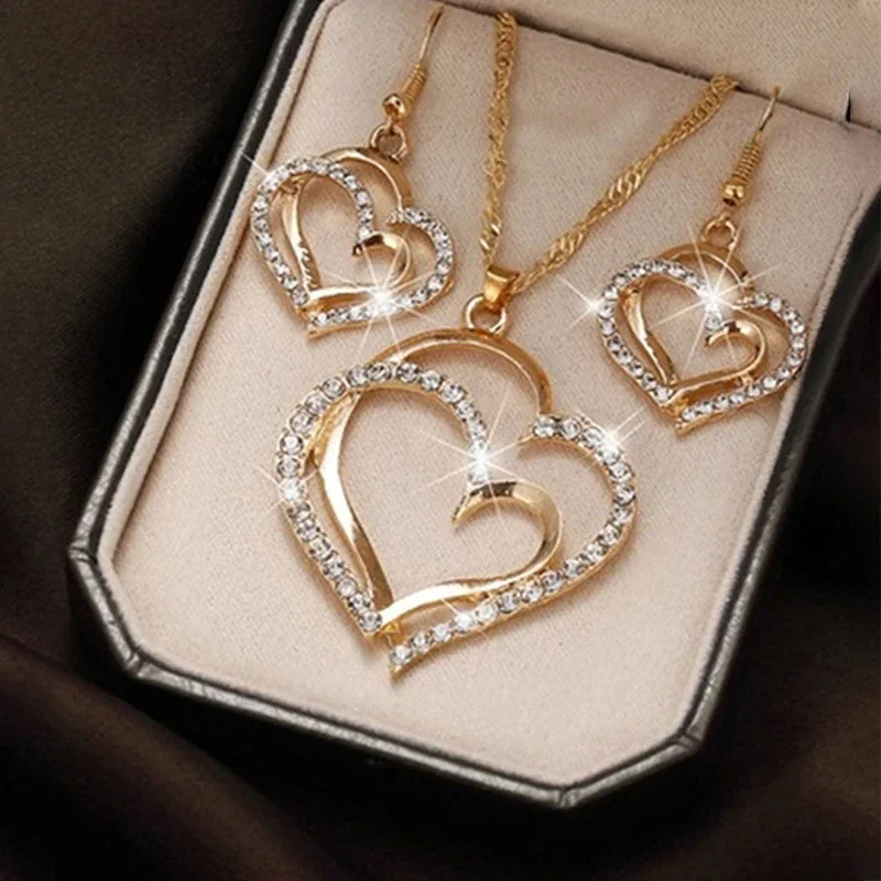 Close-up view of a gold-toned jewelry set featuring double heart pendants and earrings, adorned with sparkling crystals. The set is displayed in a beige box.