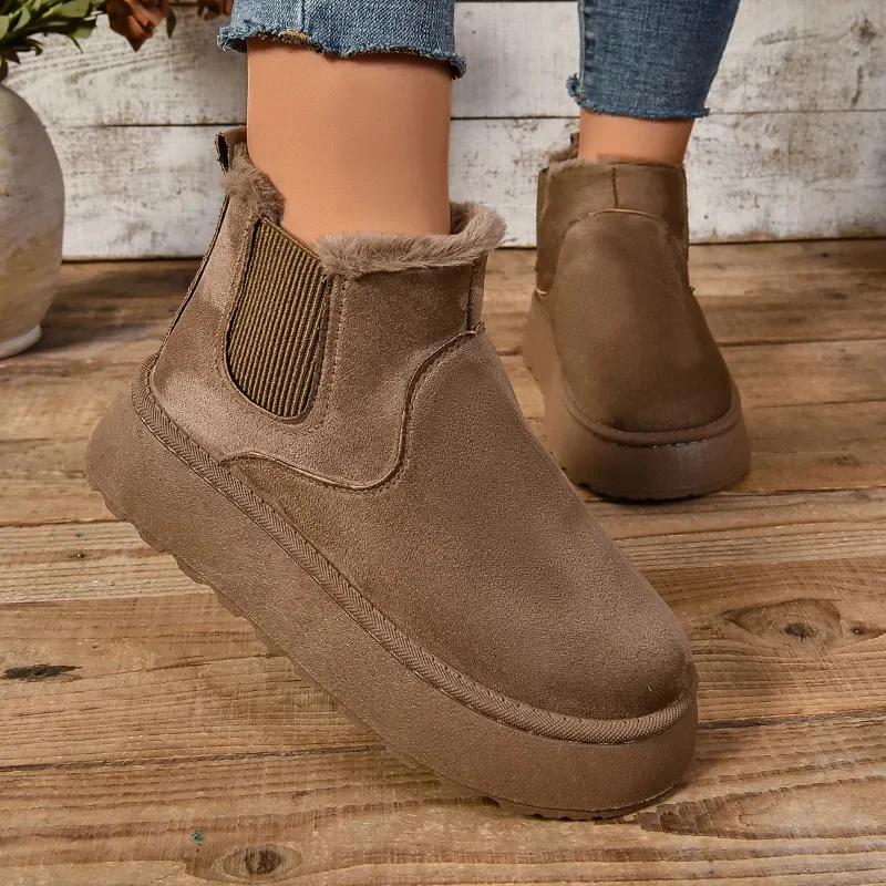 A pair of brown suede ankle boots with faux fur lining sits on a wooden surface. The boots have a chunky sole and are styled as Chelsea boots.
