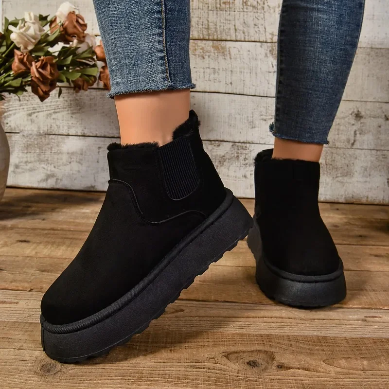 A close-up shot of a person's legs wearing black suede ankle boots with thick soles. The boots are lined with faux fur and have a slight heel. The person is wearing jeans and is standing on a wooden floor.
